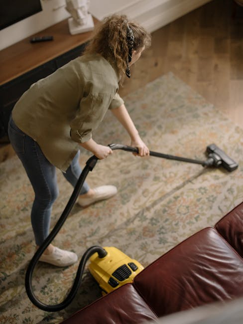 A person, dressed in black shorts and a black top, is vacuuming a beige textured carpet in a domestic setting with a red upright vacuum cleaner. The vacuum cleaner has a blue house-shaped icon on the 